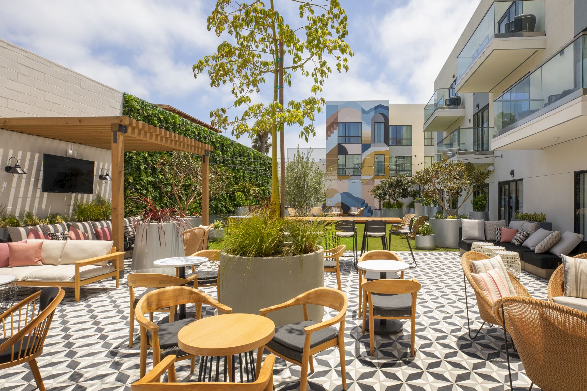 A stylish outdoor terrace with checkered tile flooring, wooden tables and chairs, planters, a shaded lounge area, and modern buildings in the background.