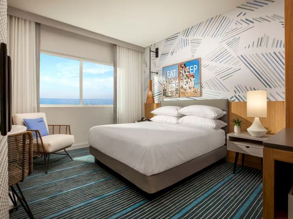 A modern hotel room with a king bed, stylish geometric wallpaper, striped carpet, two nightstands, a desk, chair, and a window view of the sea, bright and tidy.
