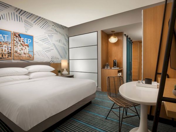 A modern hotel room with a large bed, nightstands, geometric wall art, a desk and chair, a small round table, and a doorway to a bathroom. End.