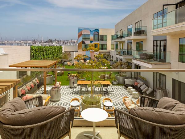 A rooftop hotel terrace with wicker chairs, cushions, and small tables, overlooking modern glass-fronted buildings and a sunny cityscape.