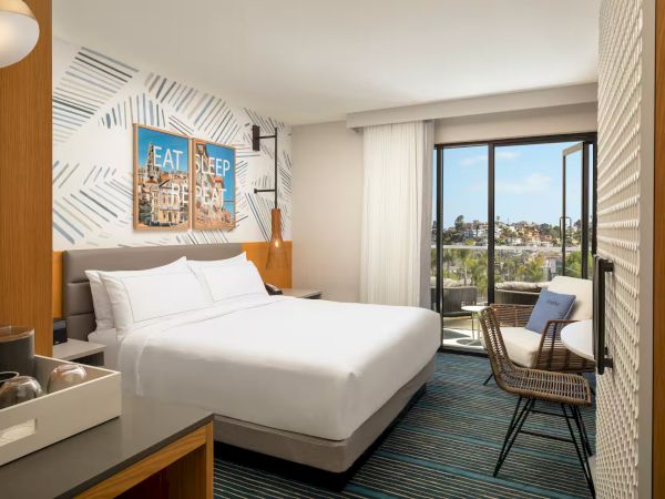 A bright hotel room with a white bed, patterned accent wall, a desk, chair, and a balcony view of a cityscape, ending with a period.