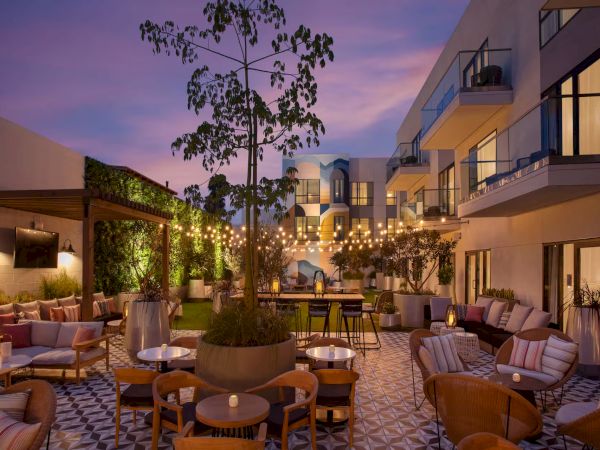 A stylish outdoor hotel courtyard at dusk with cozy seating, fairy lights, potted trees, string lights, and modern balcony rooms nearby.
