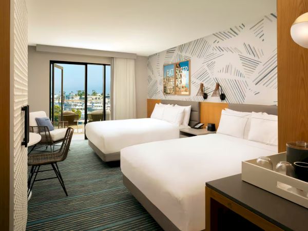 A modern hotel room with two queen beds, a seating area near a large window, and stylish geometric wall accents, ending with a view of the city.