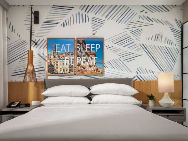 A modern hotel bedroom with a white bed, geometric blue-patterned wall, two nightstands, a lamp, and three-panel &ldquo;EAT SLEEP REPEAT&rdquo; art above the headboard, ending with a period.