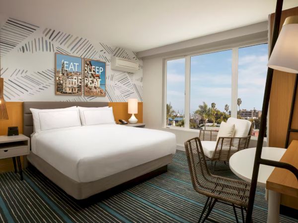 A bright hotel room with a king bed, modern headboard, patterned wall art, bedside lamps, a desk, chair, and a small table near a large window with a city/ocean view.