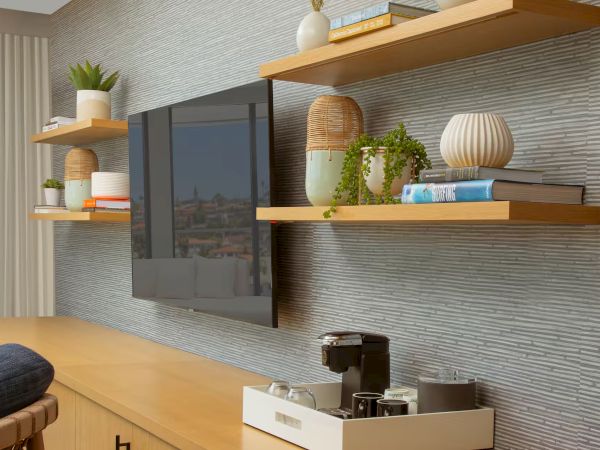 A modern living room with a mounted flat-screen TV, wooden shelves holding plants, books, and decor, plus a coffee machine on a white cart.