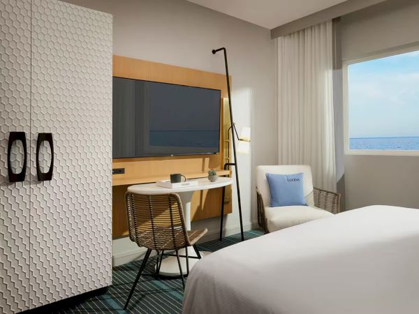 A modern hotel room with a white textured wardrobe, a desk with a mounted TV, a chair, a floor lamp, a bed, a small table, a comfy chair, and a window with an ocean view.