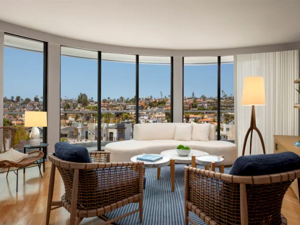 A modern, sunlit living room with curved windows, a white sofa, wicker chairs, round coffee tables, and a city view, cozy lamps, and wood flooring.