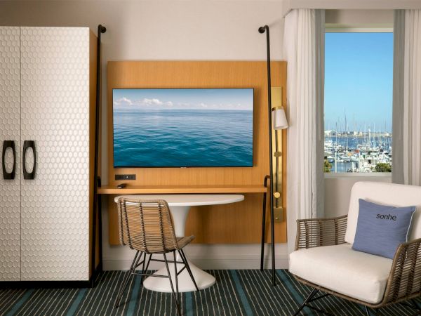 A hotel room with a TV on a wood panel, a small round table, a wire chair, a white armchair, a nearby window offering a marina view, and a tall wardrobe.