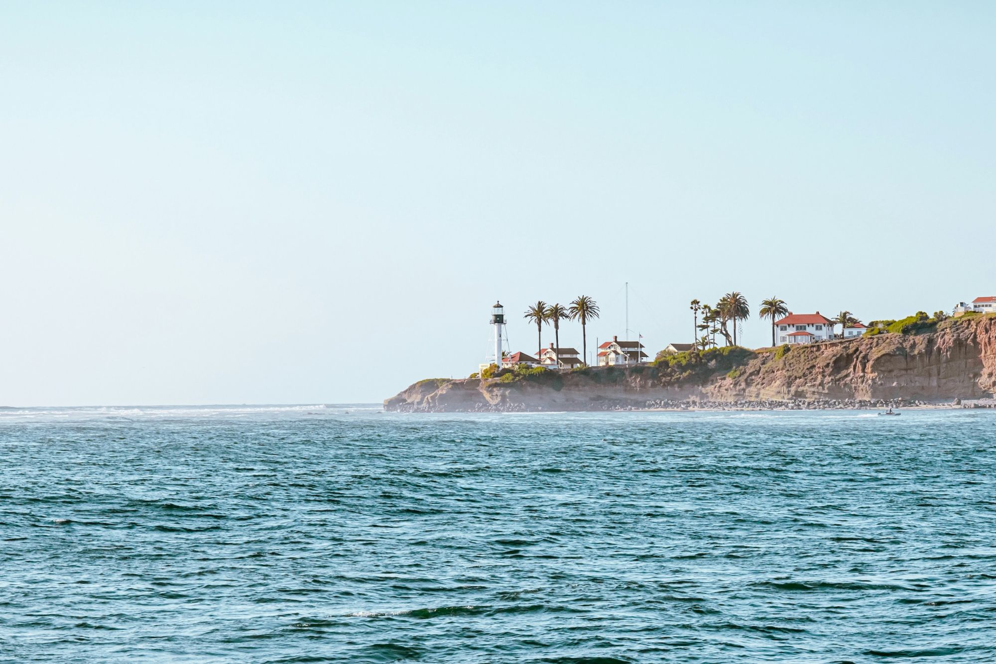 A peaceful ocean view with waves in the foreground and a rocky coastline topped with palm trees and small buildings in the distance.