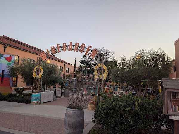 A small outdoor plaza with a decorative arch, greenery, a barrel planter, and colorful, whimsical yard art in a sunny courtyard.