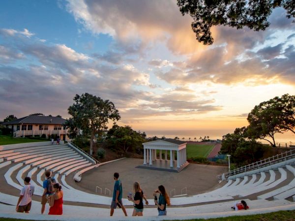 An outdoor amphitheater at sunset with curved white seating, a small white pavilion, trees, and people strolling toward the stage.