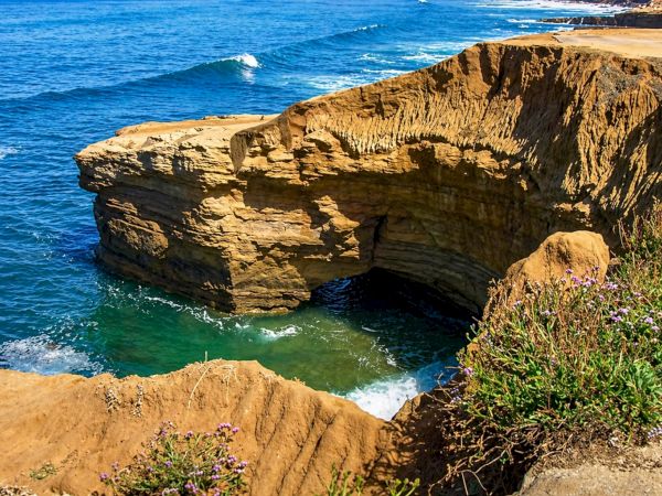 A rugged coastal cliff with a small sea cave, waves crashing against rock, and blue-green water below&mdash;sunlit and windswept.