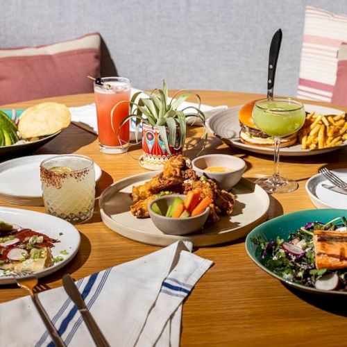 A spread of colorful dishes: salads, tacos, grilled veggies, soup, bread, chips and dips, drinks, and a cozy table setting with napkins.