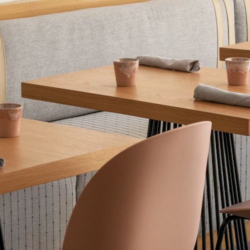 A minimalist cafe scene with wooden tables, light brown chairs, napkins, cups, and a cushioned booth in the background, ready for customers.