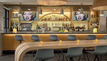 A sleek bar with a curved wooden counter, blue stools, backlit shelves of bottles, and vintage Marilyn Monroe art on the wall.