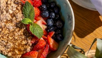 A bowl with a granola-topped yogurt or dessert, surrounded by fresh strawberries, blueberries, a mint leaf, on a wooden table near a napkin and plant, looks delicious.