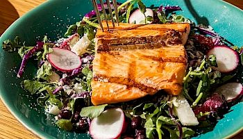 A vibrant salad with greens, radish slices, and a grilled salmon fillet, plus a pink drink in the background, all on a wooden table.