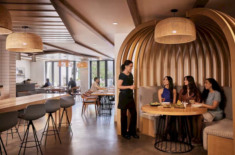 A modern restaurant or cafe with wooden, curved booths, hanging wicker lights, and patrons dining at round tables.