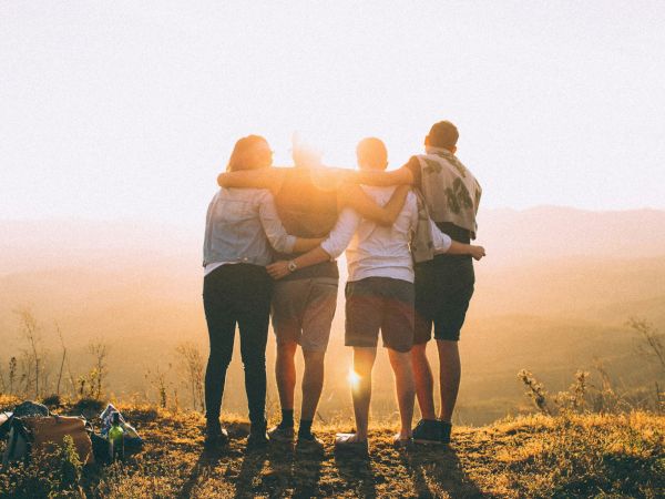 A group of five friends stands with arms around each other, watching a sunrise over a hilly landscape. Short, hopeful moment together.