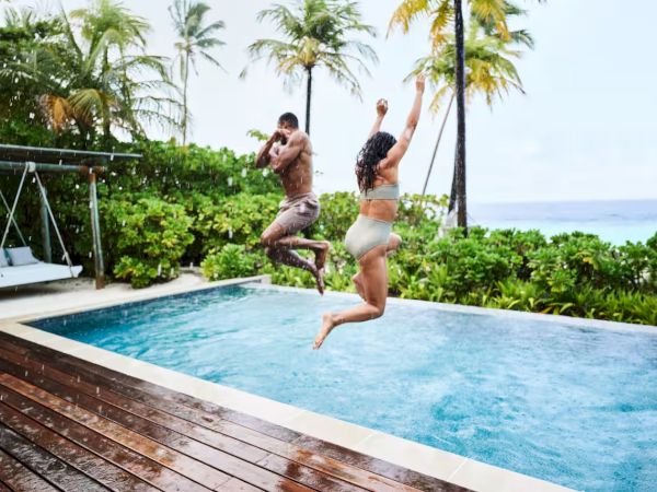 Two people are mid-air, jumping into a pool from a wooden deck by a tropical garden and palm trees, smiles on a sunny day.