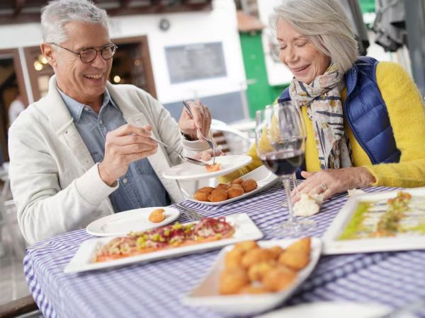Two seniors enjoying a meal at an outdoor cafe: pizza, fried appetizers, wine, and bread on a checkered tablecloth, smiling and chatting.