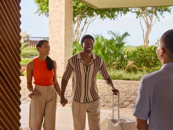 A couple greets a man with a suitcase at a sunny outdoor entrance, holding hands as they arrive at a resort lobby.