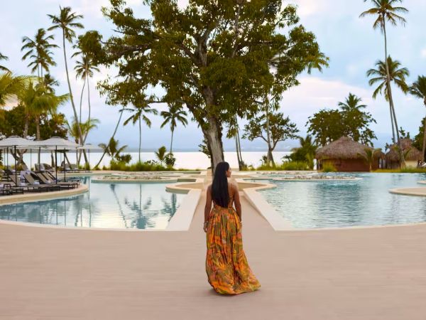A woman in a flowing orange dress walks toward a pool area at a tropical resort with palm trees and a beach in the background, sunny sky.
