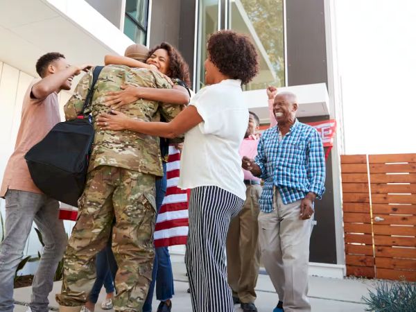 A group of people hugs a military service member at a doorway, welcoming them home; others nearby smile and cheer. Ending with a period.