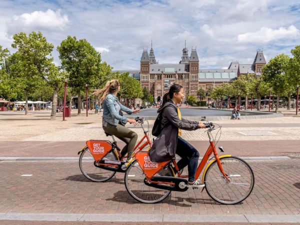 Two people ride bright orange bicycles along a city square with trees and a grand building in the background. The rider in front wears a coat.
