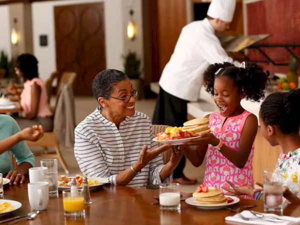 A family sharing a cheerful meal at a restaurant, kids smiling as one passes a plate to another, waiter in the background.