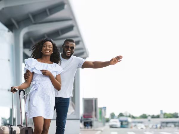 Two friends hug and smile as they arrive at the airport, one pulling a rolling suitcase, the other gesturing happily toward the road.
