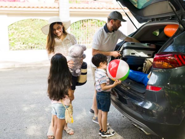 A family loads beach gear into a car: mom holds a towel and stuffed animal, kids carry a ball and towel, dad packs under a hatchback.