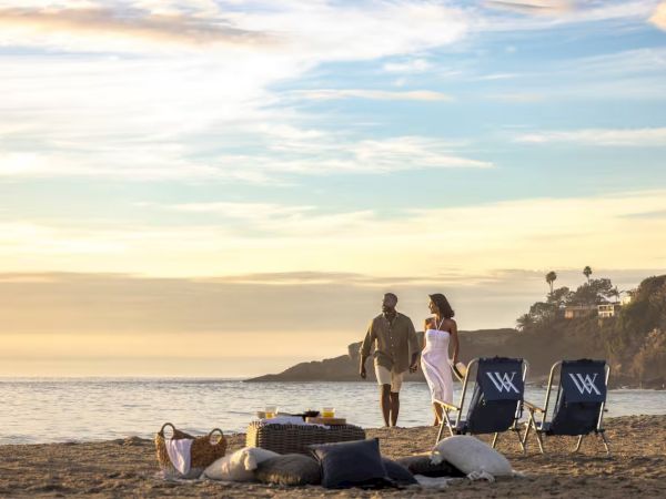 A couple walks along a sunny beach at sunset with bags, towels, and two blue beach chairs nearby. Ending the sentence.