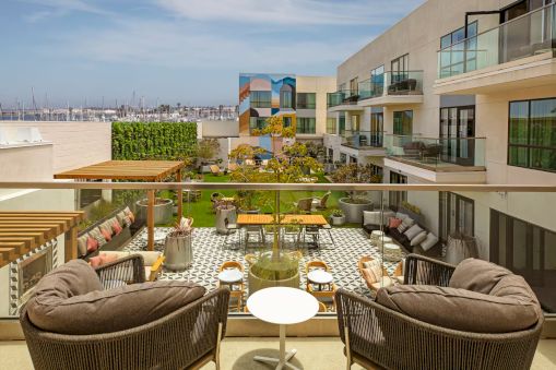 Sunny rooftop terrace with cushioned seating, small tables, and a lounge area overlooking modern apartments and blue sky, outdoors.