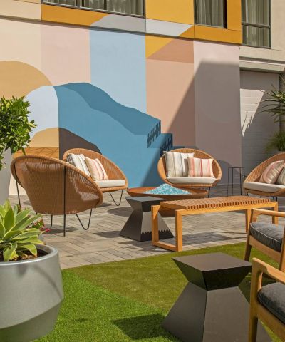 A stylish outdoor patio with wicker chairs, a wooden coffee table, potted plants, and a colorful geometric mural on the wall, sunny and inviting.