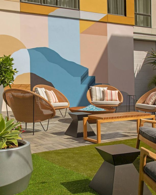 A stylish outdoor patio with wicker chairs, a wooden coffee table, potted plants, and a colorful geometric mural on the wall, sunny and inviting.