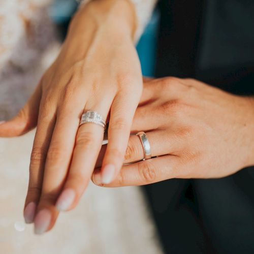 Two hands showing wedding rings, with an engagement ring and a band on the left hand, held together in a close, tender pose.