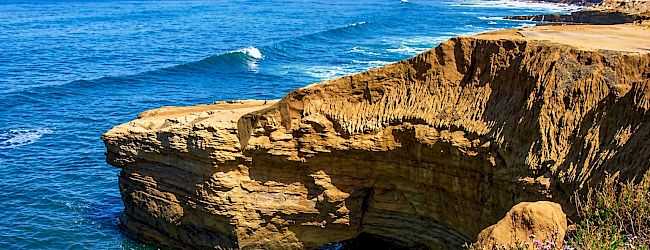 A rocky coastal cliff with turquoise water below, sunlit limestone, and a small railing at the edge.
