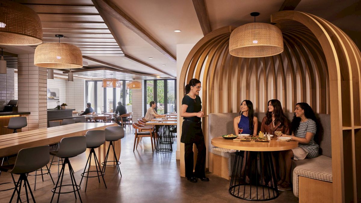 A modern, cozy restaurant with curved wooden booths, round tables, pendant lights, and a server attending to four diners at a booth.