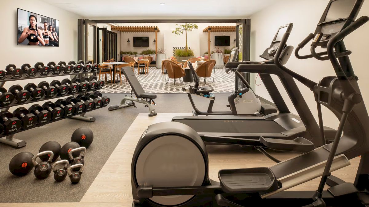 A compact home gym with treadmills, a rowing machine, weights, kettlebells, and a TV, plus a dining area visible in the background.