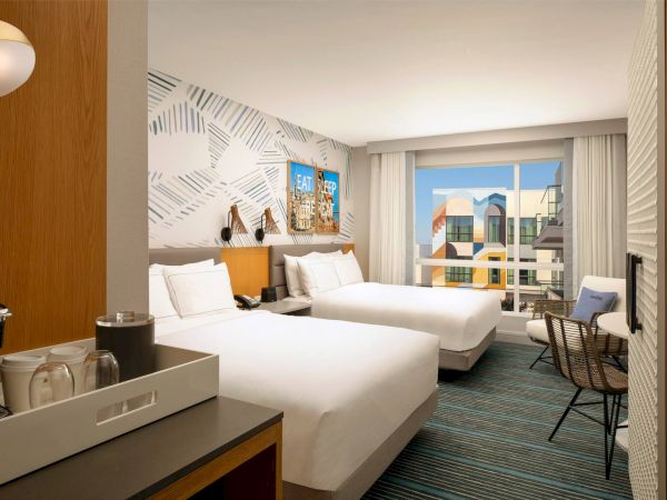 A bright hotel room with two queen beds, patterned accent wall, a desk and chairs, a window view of city buildings, and a coffee/tea setup on a tray.