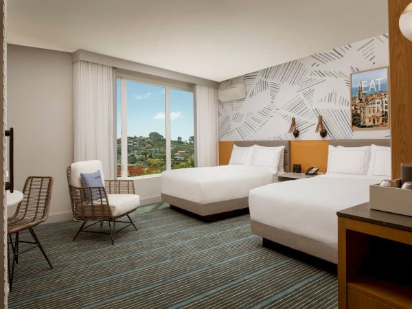 A modern hotel room with two beds, a seating area by a large window, and a stylish geometric wall pattern, bathed in natural light.