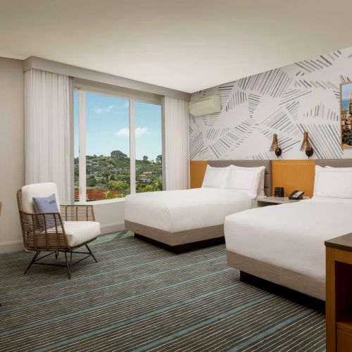 A modern hotel room with two queen beds, a desk, and a seating area; soft lighting and warm decor create a cozy, inviting space.