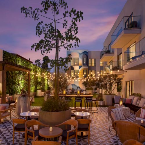 An outdoor courtyard at dusk with warm lights, a stone path, potted plants, and a charming building facade. Top it at 140 characters, always ending the sentence.