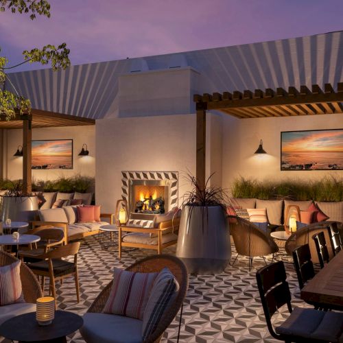 An inviting outdoor patio at dusk with: cozy seating, a lit fireplace, patterned tile floor, warm string lights, and framed art on the walls. End.