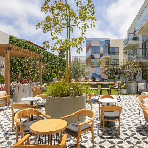A sunny outdoor patio with wooden tables and chairs, string lights, potted plants, and modern buildings in the background.
