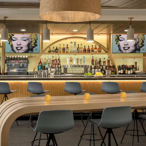 A modern bar with a curved wooden counter, row of blue chairs, shelves of liquor, and two Marilyn Monroe portraits on the back wall.