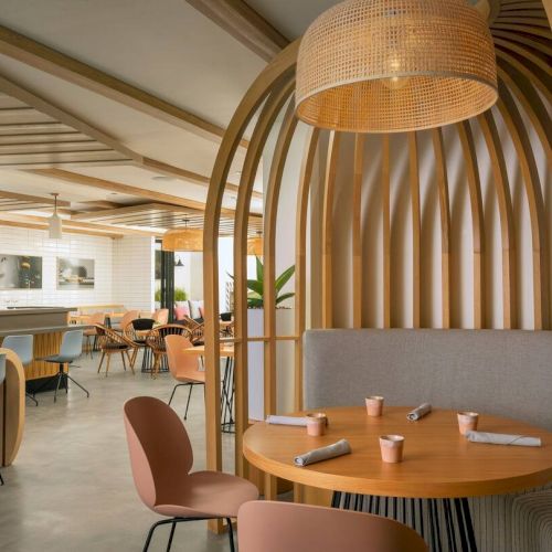 A stylish, airy cafe with curved wooden dividers, round tables, pink and gray chairs, pendant lights, and a relaxed, modern seating layout.