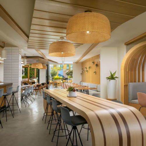 A bright, modern cafe with long wooden tables, woven pendant lights, bar seating, and cozy booths along curved wood arches, creating a welcoming atrium vibe.